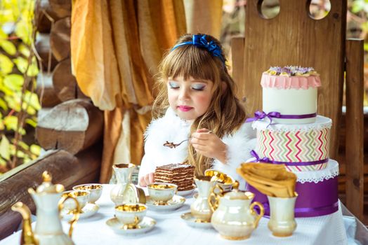 Front view of an little beautiful girl in the scenery of Alice in Wonderland holding a piece of cake on a spoon at the table in the autumn park.