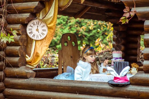 Side view of an little beautiful girl in the scenery of Alice in Wonderland holding a piece of cake on a spoon at the table in the autumn park.