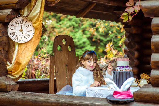 Side view of an little beautiful girl in the scenery of Alice in Wonderland holding a piece of cake on a spoon at the table in the autumn park.