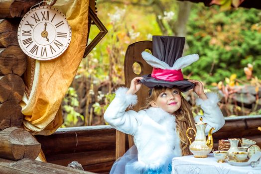 An little beautiful girl in the scenery of Alice in Wonderland holding cylinder hat with ears like a rabbit over head at the table in the garden.