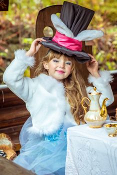 An little beautiful girl in the scenery of Alice in Wonderland holding cylinder hat with ears like a rabbit over head at the table in the garden.