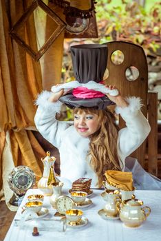 An little beautiful girl in the scenery of Alice in Wonderland holding cylinder hat with ears like a rabbit over head at the table in the garden.