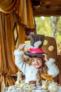 An little beautiful girl in the scenery of Alice in Wonderland holding cylinder hat with ears like a rabbit over head at the table in the garden.