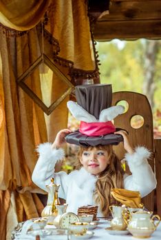 An little beautiful girl in the scenery of Alice in Wonderland holding cylinder hat with ears like a rabbit over head at the table in the garden.