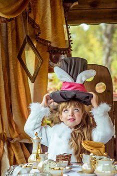 An little beautiful girl in the scenery of Alice in Wonderland holding cylinder hat with ears like a rabbit over head at the table in the garden.