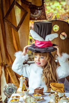 An little beautiful girl in the scenery of Alice in Wonderland holding cylinder hat with ears like a rabbit over head at the table in the garden.