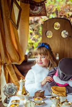 An little beautiful girl in the scenery of Alice in Wonderland holding cylinder hat with ears like a rabbit in the hands at the table in the garden.