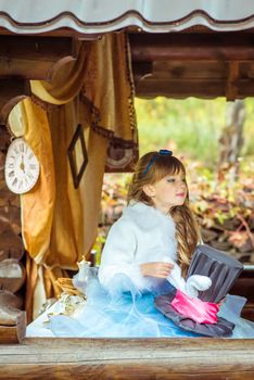 An little beautiful girl in the scenery of Alice in Wonderland holding cylinder hat with ears like a rabbit in the hands at the table in the garden.