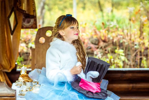 An little beautiful girl in the scenery of Alice in Wonderland holding cylinder hat with ears like a rabbit in the hands at the table in the garden.