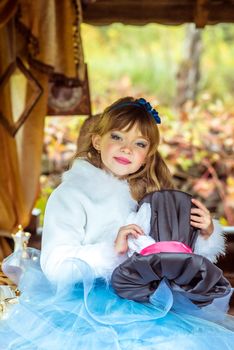 An little beautiful girl in the scenery of Alice in Wonderland holding cylinder hat with ears like a rabbit in the hands at the table in the garden.