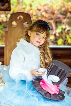 An little beautiful girl in the scenery of Alice in Wonderland holding cylinder hat with ears like a rabbit in the hands at the table in the garden.