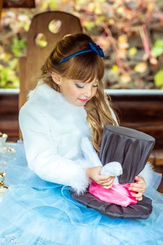 An little beautiful girl in the scenery of Alice in Wonderland holding cylinder hat with ears like a rabbit in the hands at the table in the garden.