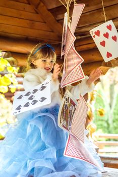 An little beautiful girl in a long blue dress in the scenery of Alice in Wonderland playing and dancing with large playing cards on the table in the garden.