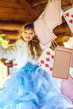 An little beautiful girl in a long blue dress in the scenery of Alice in Wonderland playing and dancing with large playing cards on the table in the garden.