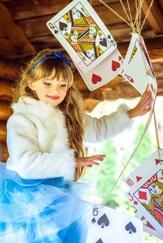 An little beautiful girl in a long blue dress in the scenery of Alice in Wonderland playing and dancing with large playing cards on the table in the garden.