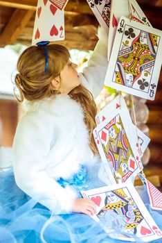 An little beautiful girl in a long blue dress in the scenery of Alice in Wonderland playing and dancing with large playing cards on the table in the garden.