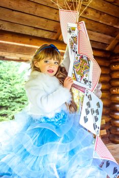 An little beautiful girl in a long blue dress in the scenery of Alice in Wonderland playing and dancing with large playing cards on the table in the garden.