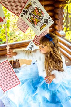 An little beautiful girl in a long blue dress in the scenery of Alice in Wonderland playing and dancing with large playing cards on the table in the garden.