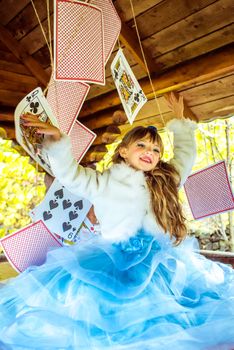 An little beautiful girl in a long blue dress in the scenery of Alice in Wonderland playing and dancing with large playing cards on the table in the garden.