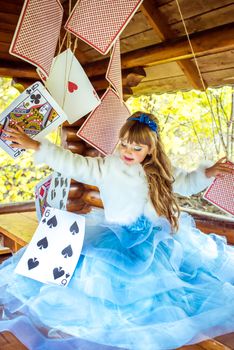 An little beautiful girl in a long blue dress in the scenery of Alice in Wonderland playing and dancing with large playing cards on the table in the garden.