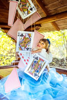 An little beautiful girl in a long blue dress in the scenery of Alice in Wonderland playing and dancing with large playing cards on the table in the garden.