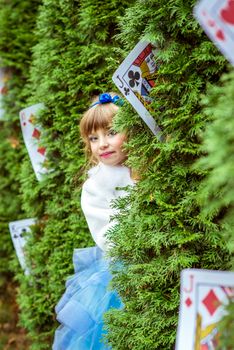 An little beautiful girl in a long blue dress in the scenery of Alice in Wonderland looking from under the fir trees are decorated with large playing cards.