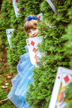 An little beautiful girl in a long blue dress in the scenery of Alice in Wonderland looking from under the fir trees are decorated with large playing cards.