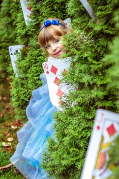 An little beautiful girl in a long blue dress in the scenery of Alice in Wonderland looking from under the fir trees are decorated with large playing cards.