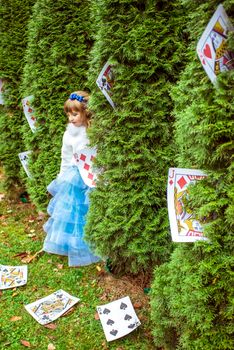 An little beautiful girl in a long blue dress in the scenery of Alice in Wonderland standing near the fir trees and looking down at the playing cards.