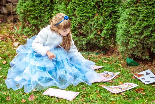 An little beautiful girl in a long blue dress in the scenery of Alice in Wonderland sitting on the grass and playing with large game cards.