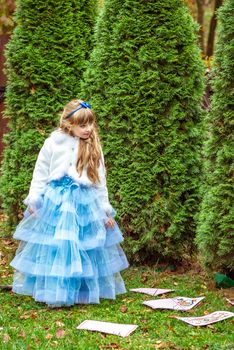 An little beautiful girl in a long blue dress in the scenery of Alice in Wonderland standing near the fir trees and looking down at the playing cards.