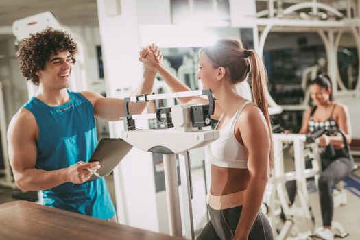 Fitness instructor with smiling sporty girl measuring her weight in gym.