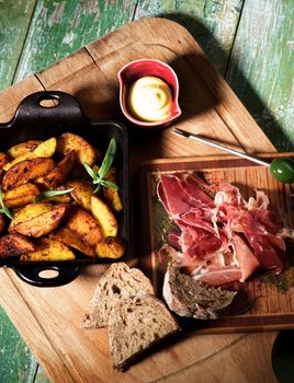 Arrangement of Cured Jamon with Grey Bread and Roasted Potato Wedges with Rosemary in Black Cast-Iron Pan with Cheese Sauce closeup on Wooden Board in Shadow