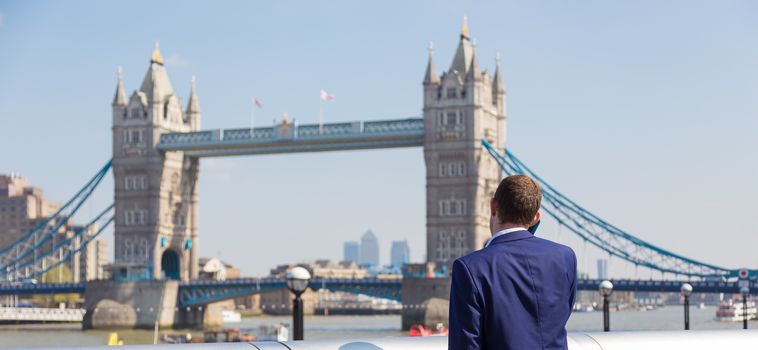 Businessman talking on mobile phone outdoor, looking at Tower Bridge in London city, UK.