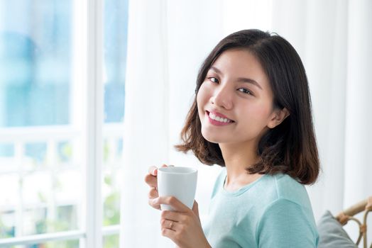 Young pretty woman sitting at opened window drinking coffee and looking outside enjoys of rest