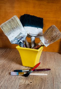 Old large paint brushes stand in a yellow vase and next to them on a wooden board lie small brushes