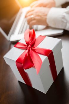 White Gift Box with Red Ribbon and Bow Near Man Typing on Laptop Computer.