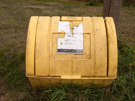 a yellow grit box outside with its lid closed and on the grass next to a path winter roads salt roads and cars accidents prevention