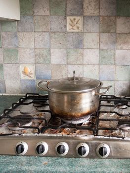 A Big Silver Metal Pan with Lid on And with A Burnt black and Brown Bottom Base Resting on Top of Gas Cooker hob with Dials all Turned off, and with bits of dirt and burnt food on some foil underneath the metal frame and with tiles in the background in UK Kitchen Unclean