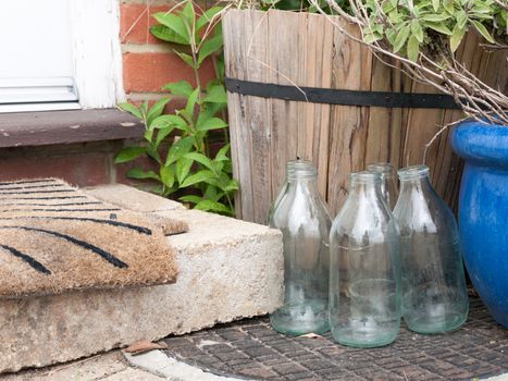 close up cool and modern shot of some glass milk bottles glasses outside empty no milk next to front door step and matt dairy retro vintage food ingredients drink england uk milkman delivery no bottle tops