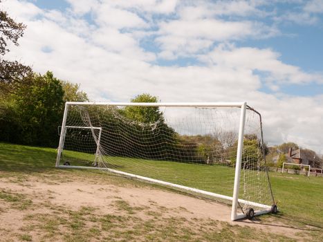 An Empty and Unused Goal Post with A White Net in the Middle of A Park with Grass and Soil on the Ground, Wheels on the Frame to Help Move it and Houses and Trees in the Background, Used For Football and for A Goal Keeper No Person in the Spring in the UK