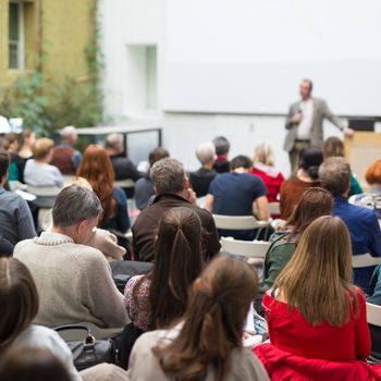 Male speaker giving presentation in lecture hall at university workshop. Audience in conference hall. Rear view of unrecognized participant. Scientific conference event. Copy space on whitescreen.