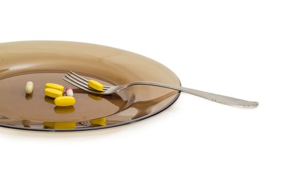 Fragment of the big dark glass dish and a stainless steel fork with several yellow tablets of the multivitamins and different dietary supplements in the form of capsules closeup on a light background
