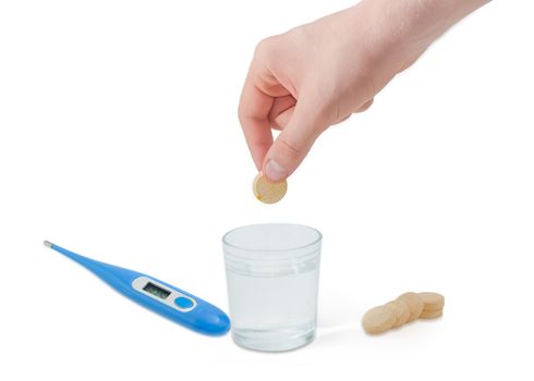 Effervescent medicinal tablet in the human hand above a glass of water, several same tablets beside and blue electronic clinical thermometeron a light background
