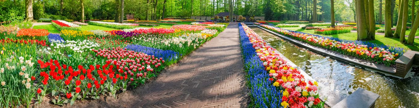 Panorama of the part of the garden with flower beds with tulips and other flowers beside to a small channel in Keukenhof Park
