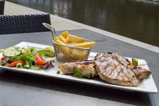 Plate of grilled meats with fries and salad on a table along the Moselle