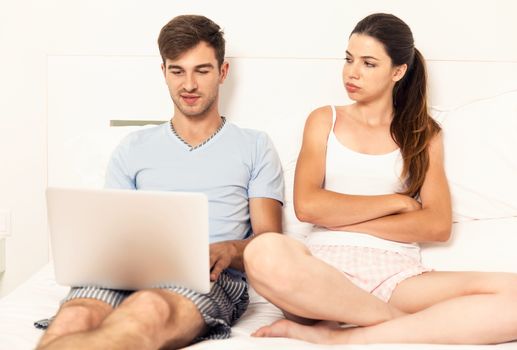 Addicted young man on bed with a laptop while the woman looks angry