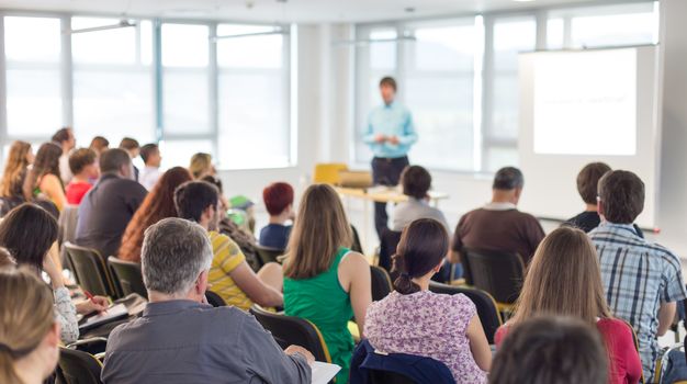 Business and entrepreneurship symposium. Speaker giving a talk at business meeting. Audience in conference hall. Rear view of unrecognized participant in audience.