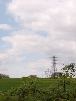 a shot of far away field green and with several lined up electrical wire metal frames standing and with a beautiful background of white and blue skies during the day of spring and trees upfront near traintracks