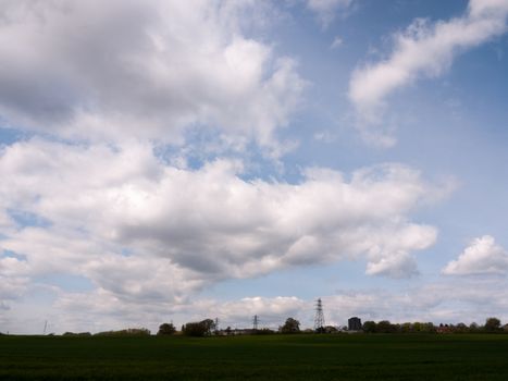 a shot of far away field green and with several lined up electrical wire metal frames standing and with a beautiful background of white and blue skies during the day of spring and trees upfront near traintracks stunning